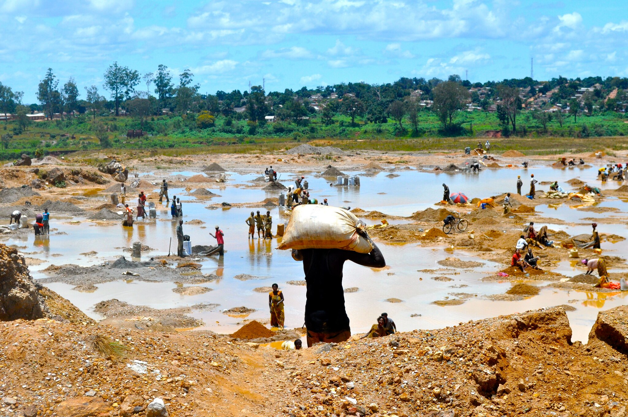 Zahlreiche Menschen arbeiten unter freiem Himmel im Kupferabbau in einer schlammigen, wasserbedeckten Landschaft im Kongo. Eine Person trägt einen schweren Sack auf dem Rücken, während andere in kleinen Gruppen graben oder schürfen. Im Hintergrund ist eine grüne Vegetation und eine Siedlung zu sehen.