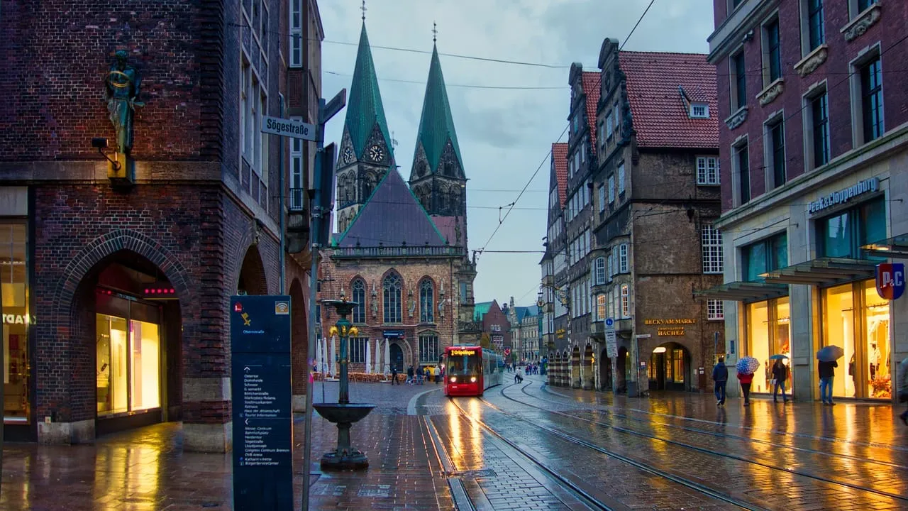 Abendstimmung in der Bremer Altstadt: Blick auf den St.-Petri-Dom mit seinen markanten Doppeltürmen. Eine Straßenbahn fährt über das nasse Kopfsteinpflaster, während Passanten mit Regenschirmen an den beleuchteten Geschäften entlanggehen.