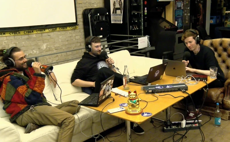 Rainer, Benjamin und Konrad mit Mikrofonen in den Händen in den Räumen des Chaos Computer Club Berlin. Auf dem Tisch Kabel, Laptops, eine Winkekatze. Im Hintergrund eine unverputzte Mauer, Arcade-Automaten, ein Mateautomat, eine riesige Leinwand und gelb schwarzes Cyber-Absperrband.